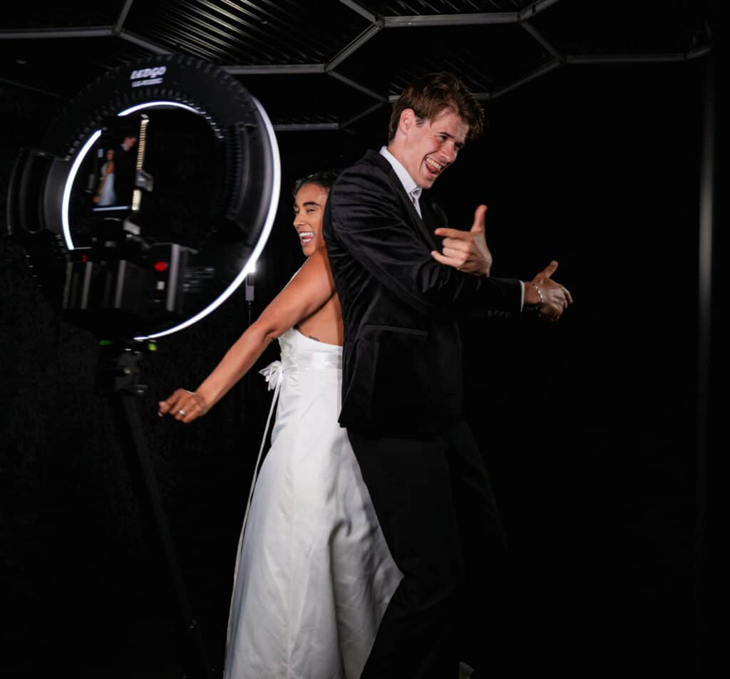 A bride and groom stand back-to-back, smiling and making playful hand gestures in front of a photo booth rental setup with a ring light and camera against a dark background.