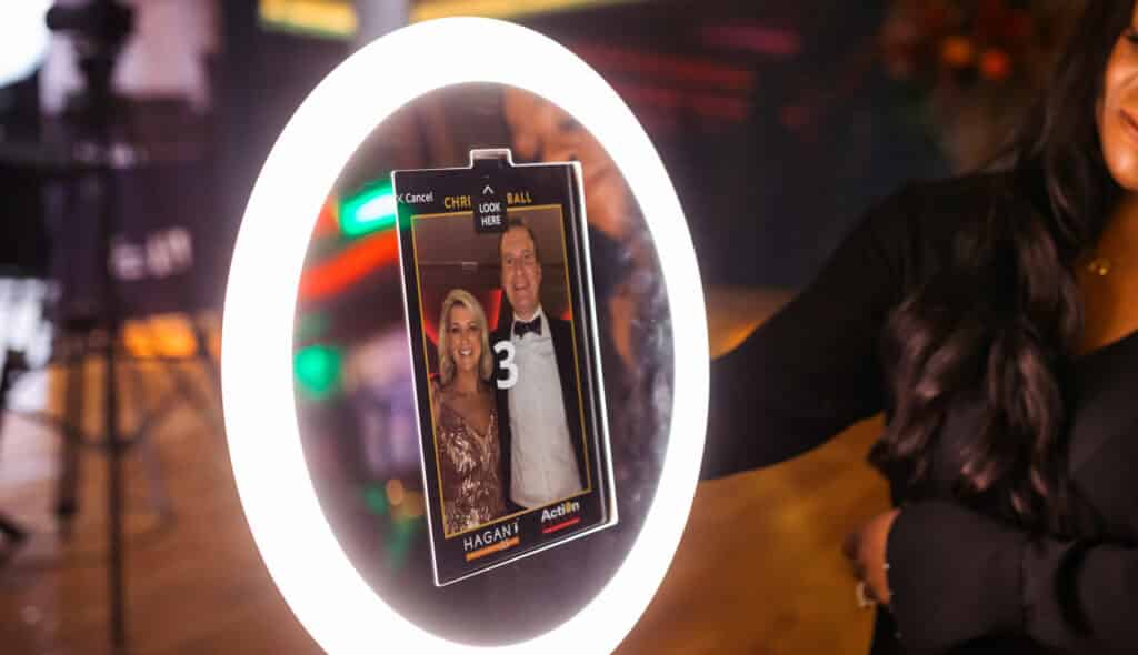 A woman uses a photo booth rental with a ring light. The screen displays a countdown and a preview photo of a smiling man and woman dressed in formal attire.