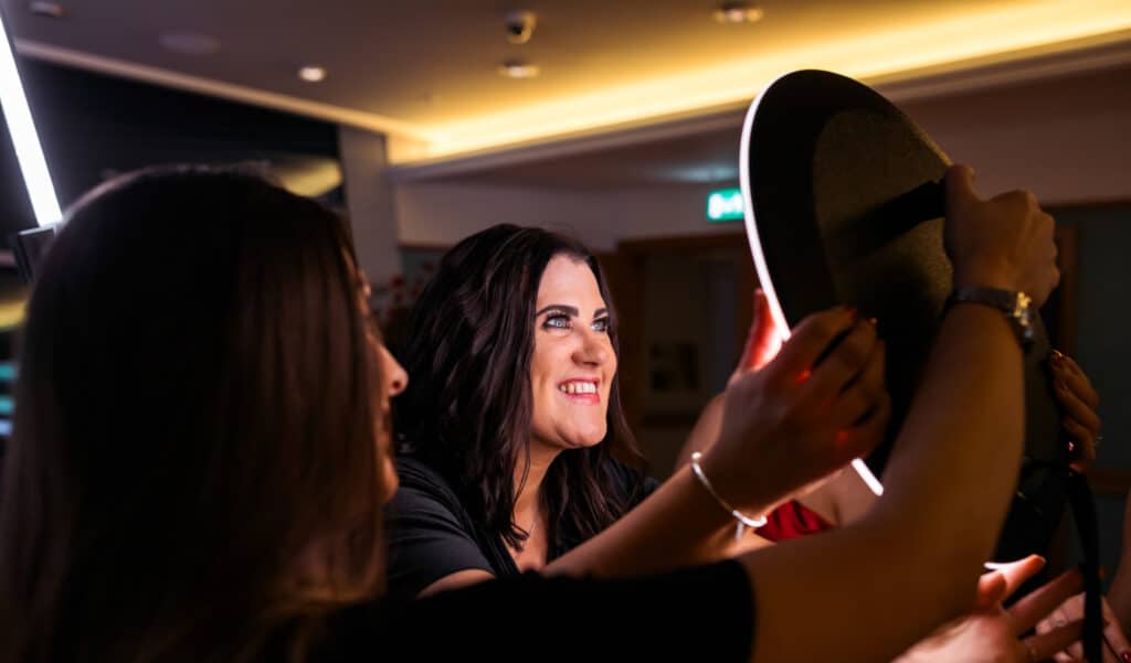 Two women smile and adjust a circular ring light in a warmly lit indoor setting, creating a bright, cheerful atmosphere—perfect inspiration for your next photo booth rental.