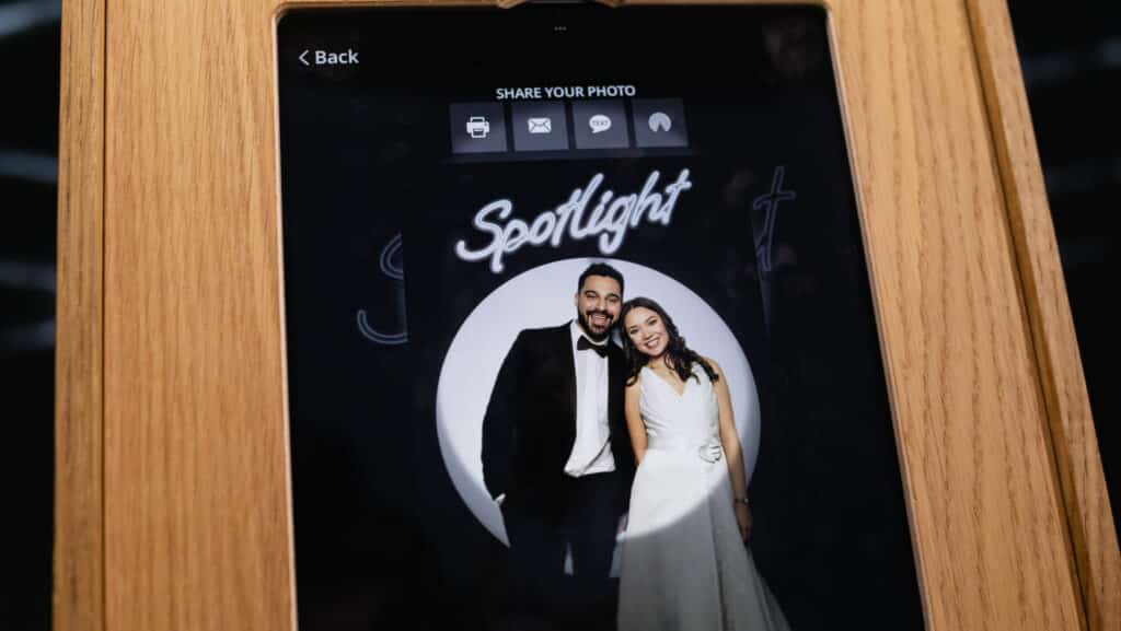A tablet screen displays a photo of a smiling man in a tuxedo and a woman in a white dress standing before a “Spotlight” backdrop, taken with a photo booth rental. Sharing options are visible at the top of the screen.
