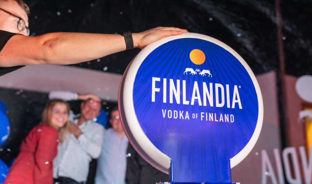 A person presses a large blue Finlandia Vodka button while three smiling people pose in the background at a photo booth rental event. The scene is lively with festive lighting and decorations.