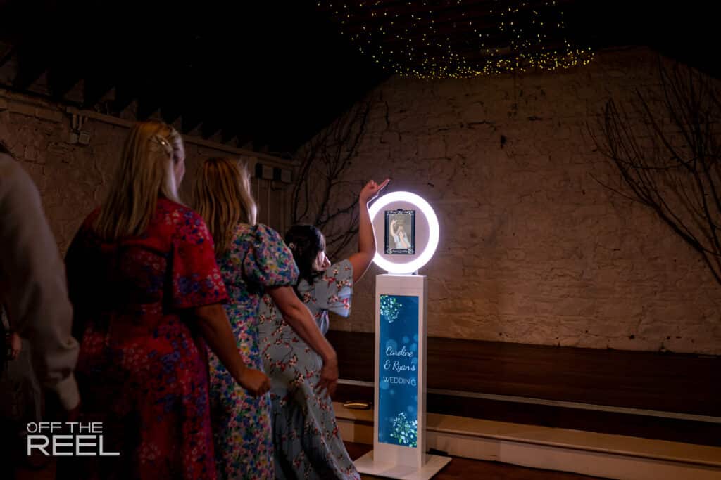 Three women in dresses facing a glowing ring light photo booth setup in  a dark room